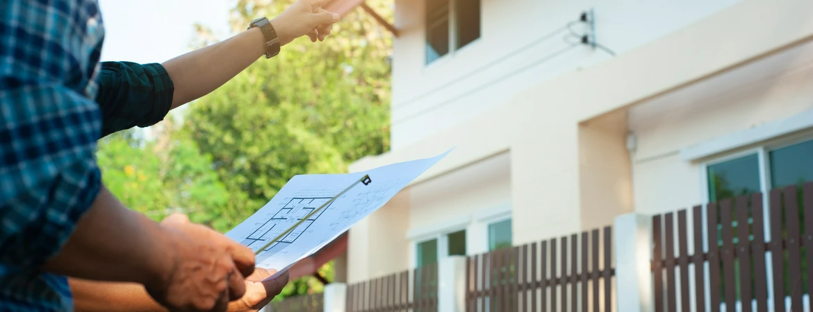 Zbliżenie na&nbsp;ręce osoby trzymającej projekt na&nbsp;tle nowoczesnego, białego domu budowlany