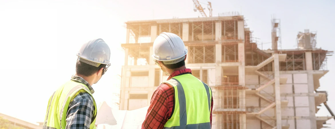 Dwóch inżynierów budowy patrzy na&nbsp;powstający wielopiętrowy budynek o&nbsp;konstrukcji żelbetowej
